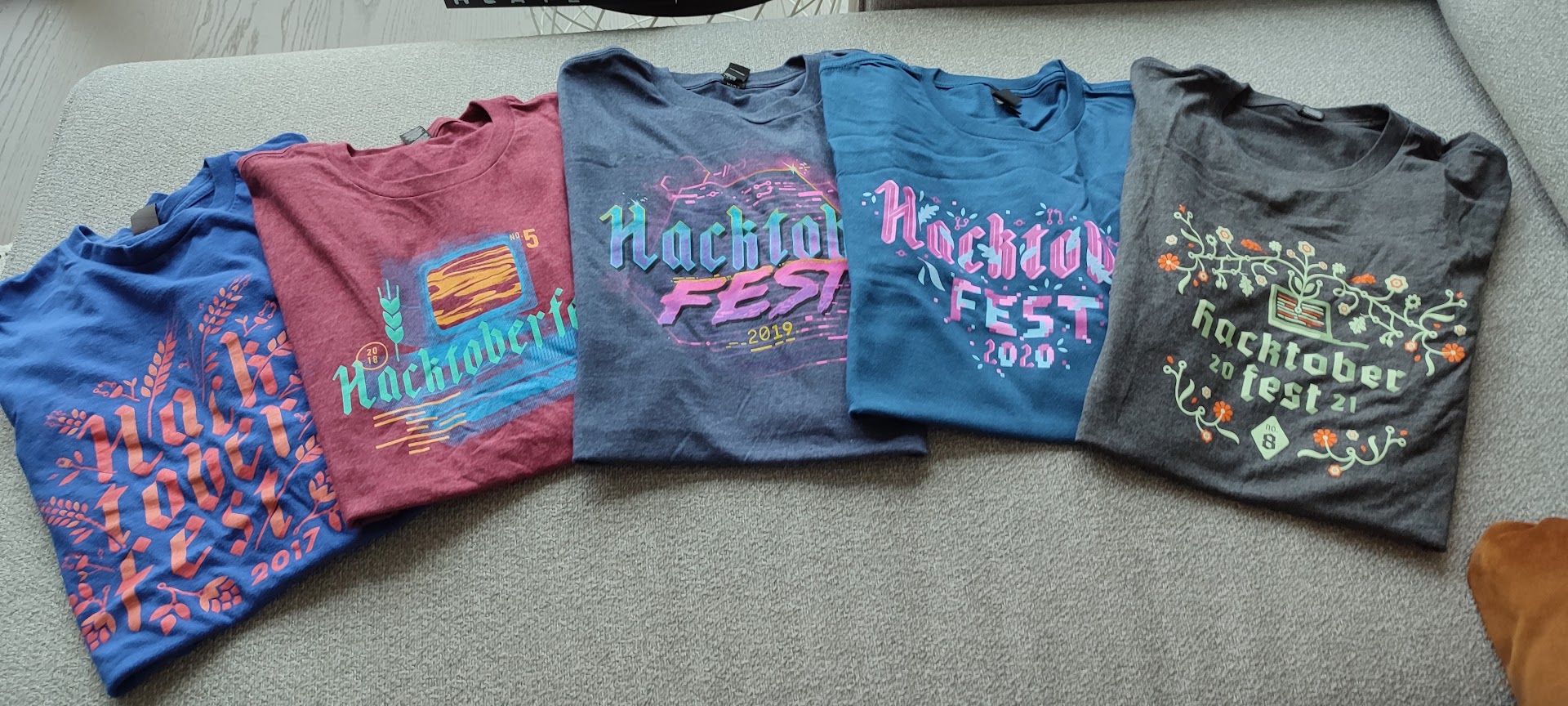 Five Hacktoberfest t-shirts, dated 2017 through 2021, laid out in a row on a light grey carpet.