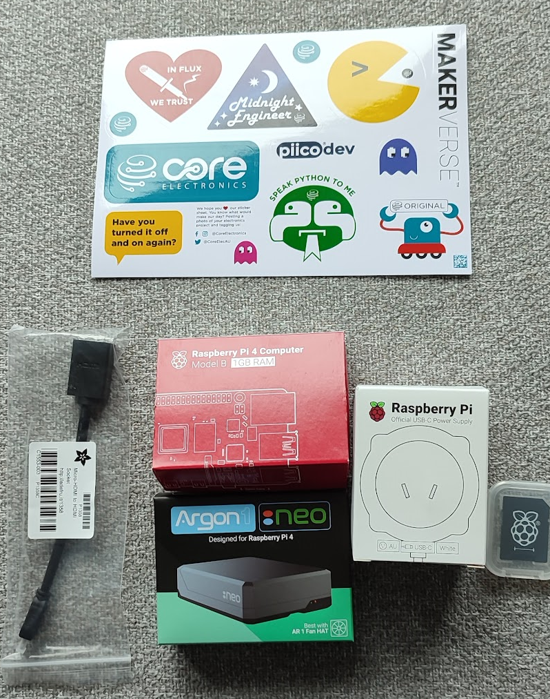 A photograph on a grey carpet of the parts: a sheet of Core Electronics stickers, a USB cable in a plastic bag, a red Raspberry Pi 4 Model B box, a black Argon NEO case box and a small microSD card adapter.