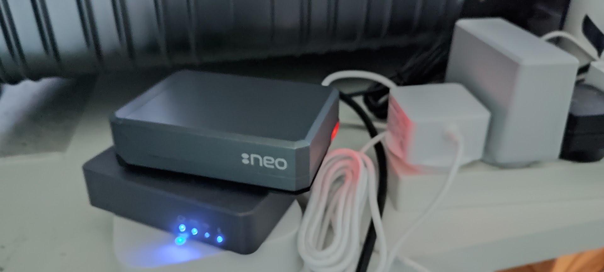 A photograph of the Raspberry Pi in its black case on a desk, powered up with a blue status light, beside a tangle of cables and a white plug.